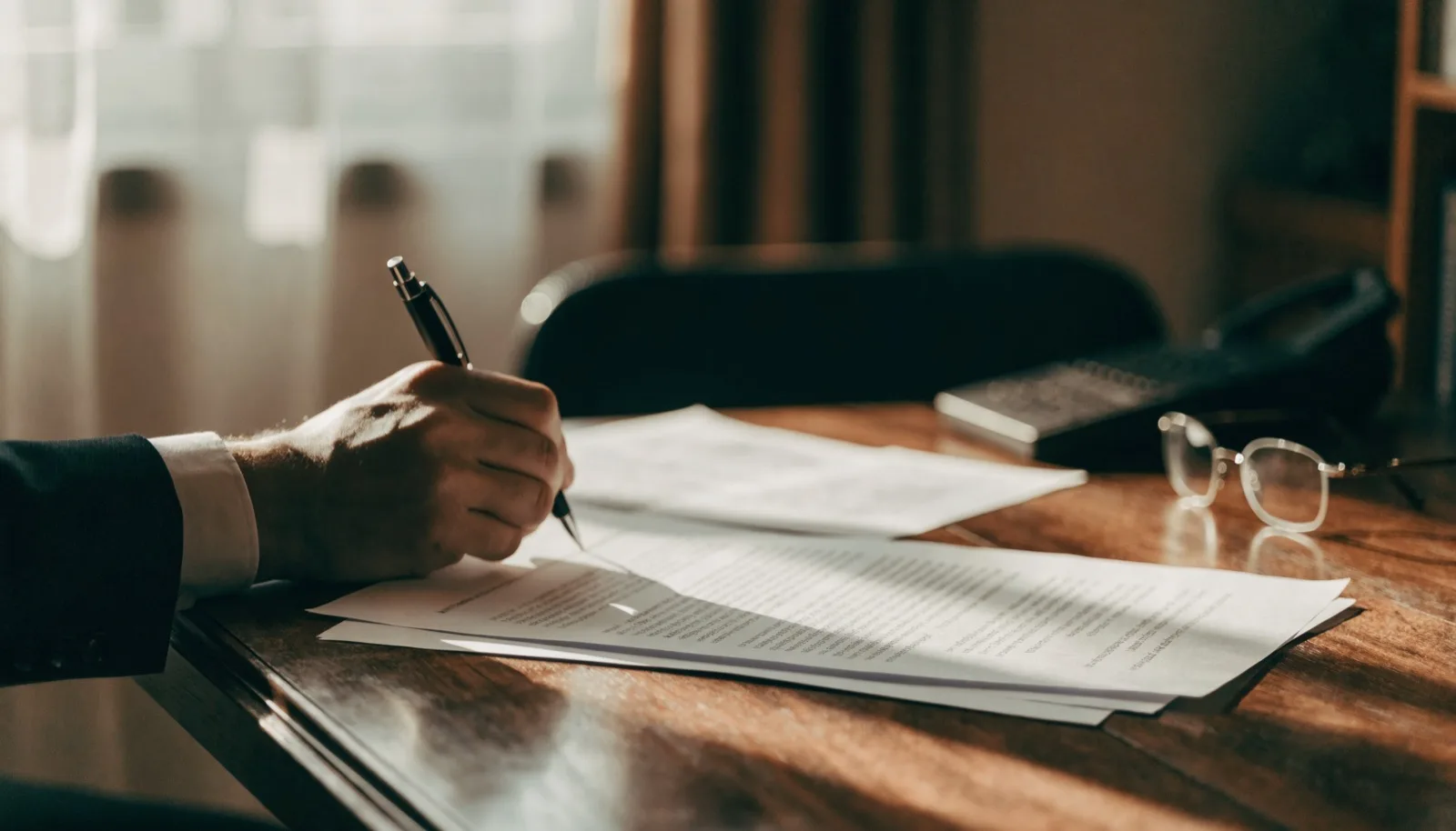 Mano che firma un contratto d'affitto su una scrivania in legno, occhiali da lettura accanto